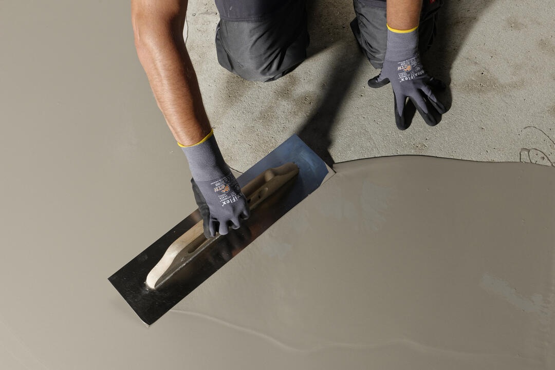 ARDEX K 15 N-Spachtelmasse wird von einer knienden Person mit einem Spachtel auf dem Boden verteilt. Sie hat einen satt-warmen, hellgrauen Farbton.