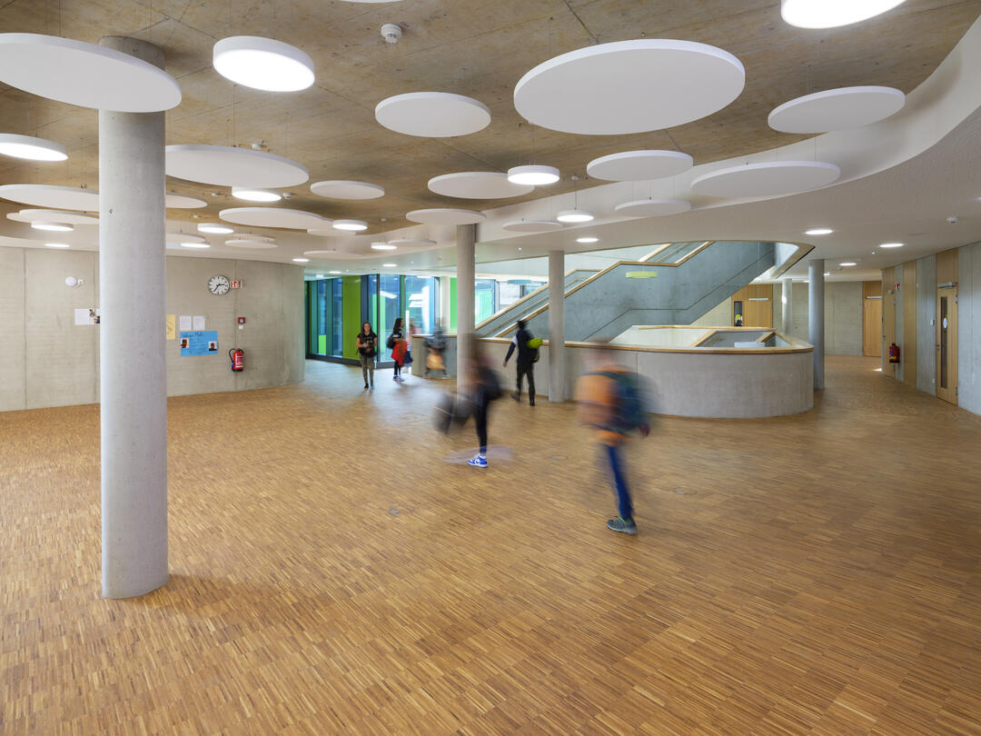 Ein minimalistischer, weitläufiger Eingangsbereich zu einer Schule mit Dielenboden und runden Rockfon-Elementen an der Decke, die in etwas größerem Abstand montiert sind.