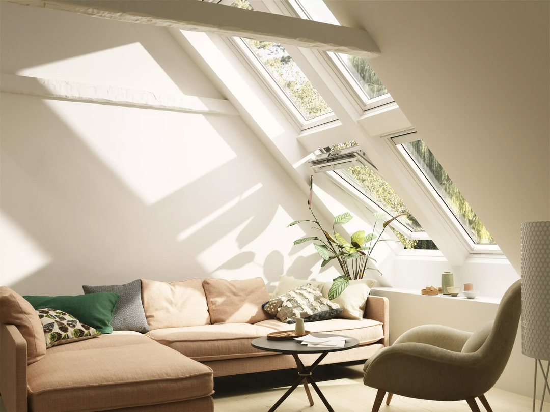 Ein lichtdurchflutetes Wohnzimmer unter einer Dachschräge mit Velux-Fenstern. Der Raum ist modern eingerichtet und die vorherrschende Farbe ist beige.
