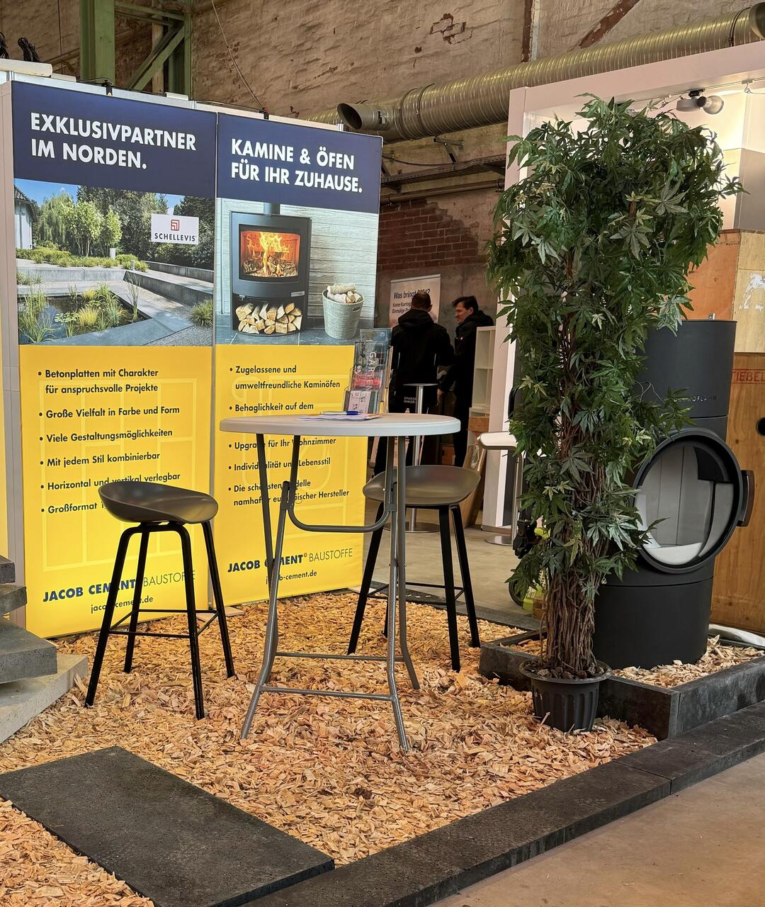 Ein mit Holzsplitt ausgestreuter Messestand mit einer Sitzgelegenheit und JACOB CEMENT-Aufstellern und einer Grünpflanze im Hintergrund.