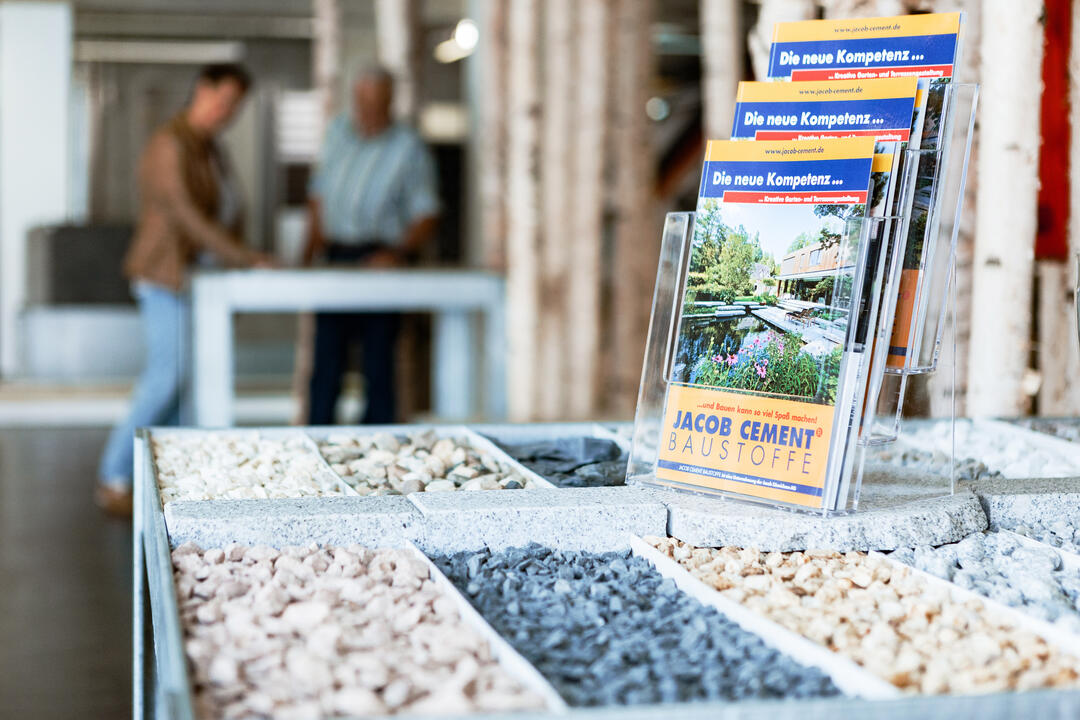 In kleinen Einbuchtungen gelagerte Gartenbaustoffe im Innenraum einer JACOB-CEMENT-Ausstellung, daneben JACOB CEMENT-Prospekte.