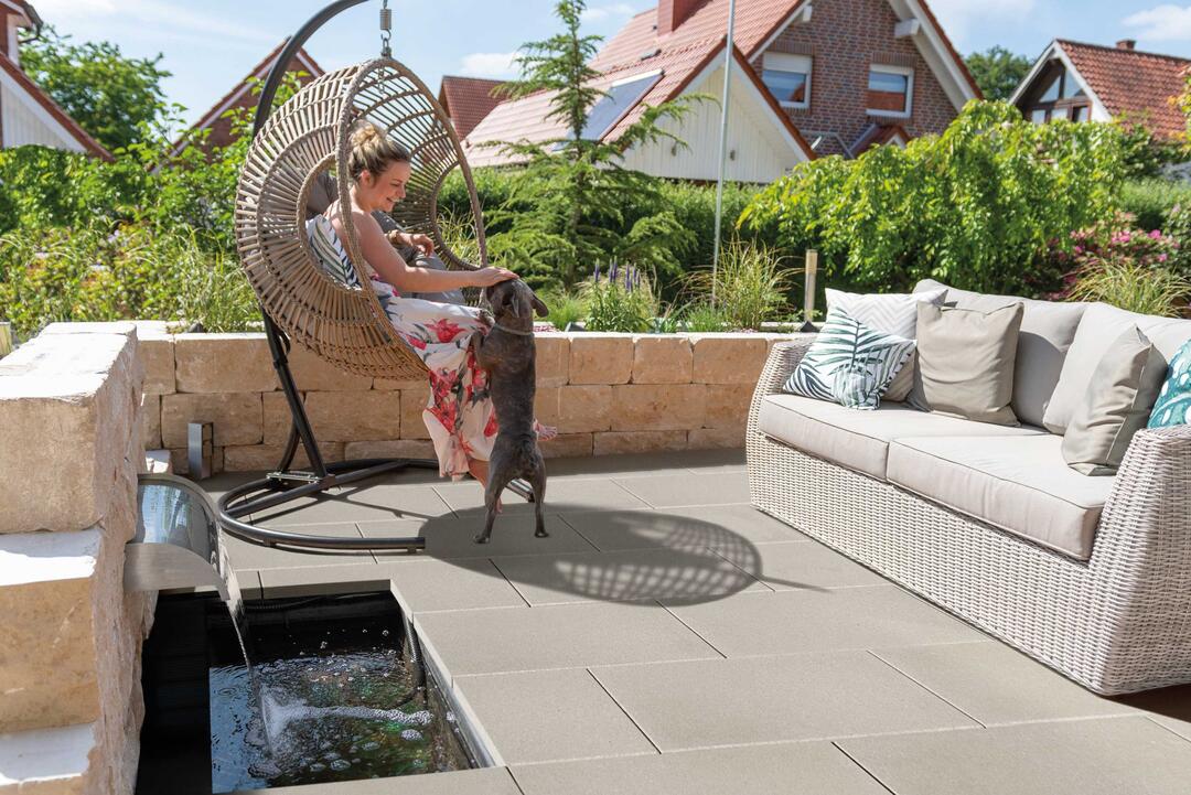 Ambientebild von einer Terrasse aus LuxoGran-Terrassenplatten in der Farbvariante Sand, auf der ein Sofa steht. In einem Hängesessel sitzt eine lachende Frau, die mit einem kleinen Hund spielt.
