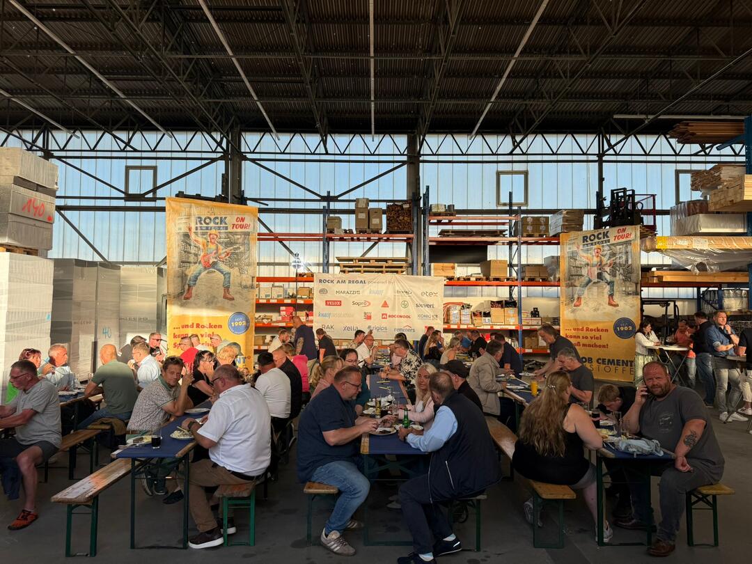 Bei einer ROCK-AM-REGAL-Veranstaltung von JACOB CEMENT sitzen Menschen an Bierzeltgarnituren in einer Lagerhalle.