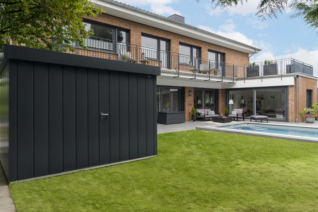 Ein Garten vor einem Haus, im Vordergrund ein schwarzes, quadratisches Gerätehaus, modern und schnörkellos.