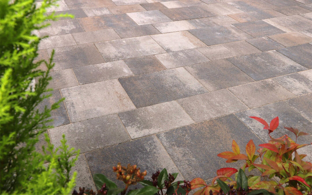 Blick von schräg oben auf eine gepflasterte Fläche, die von blühenden Pflanzen umgeben ist. Das braun-grau-rötliche Pflaster wirkt rustikal und beständig.