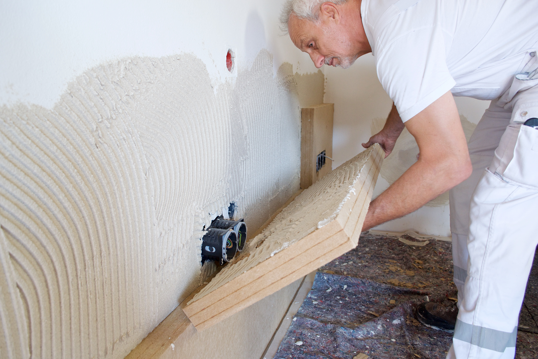 Ein Handwerker in weißer Arbeitskleidung bringt eine Platte der STEICO-Innendämmung an einer Wand an. Hier bückt er sich und sieht sich die Position genau an.