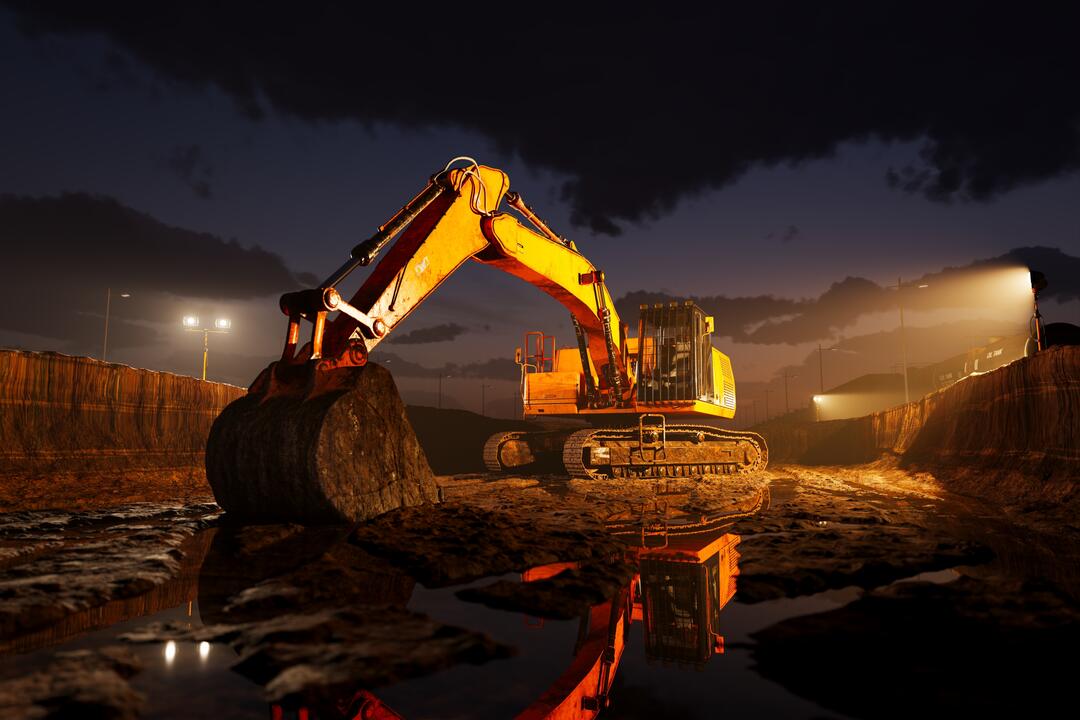 Ein gelber Bagger steht ruhend auf einer nächtlichen Baustelle, die von gelblichem Licht indirekt erleuchtet wird.