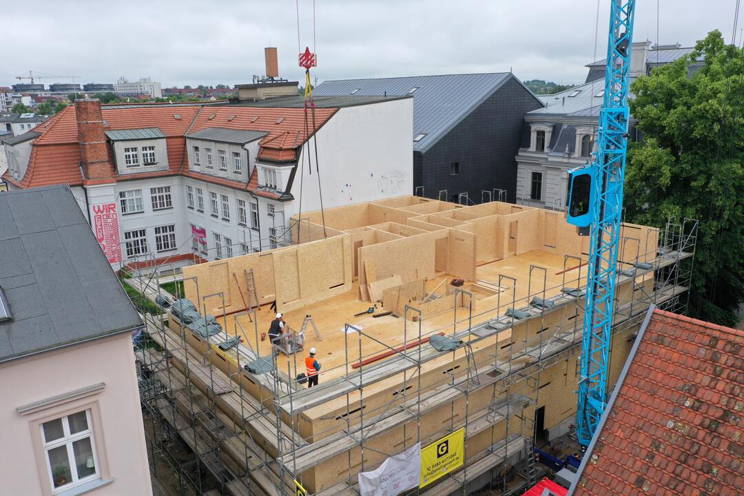 Eine Baustelle oben auf einem Dach, in Holzbauweise, mit einem blauen Kran daneben und Stadt im Hintergrund.