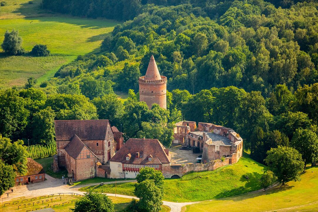 Die Burg in Burg Stargard in Neubrandenburg, rötlich inmitten dichten grünen Walds und Wiesen.