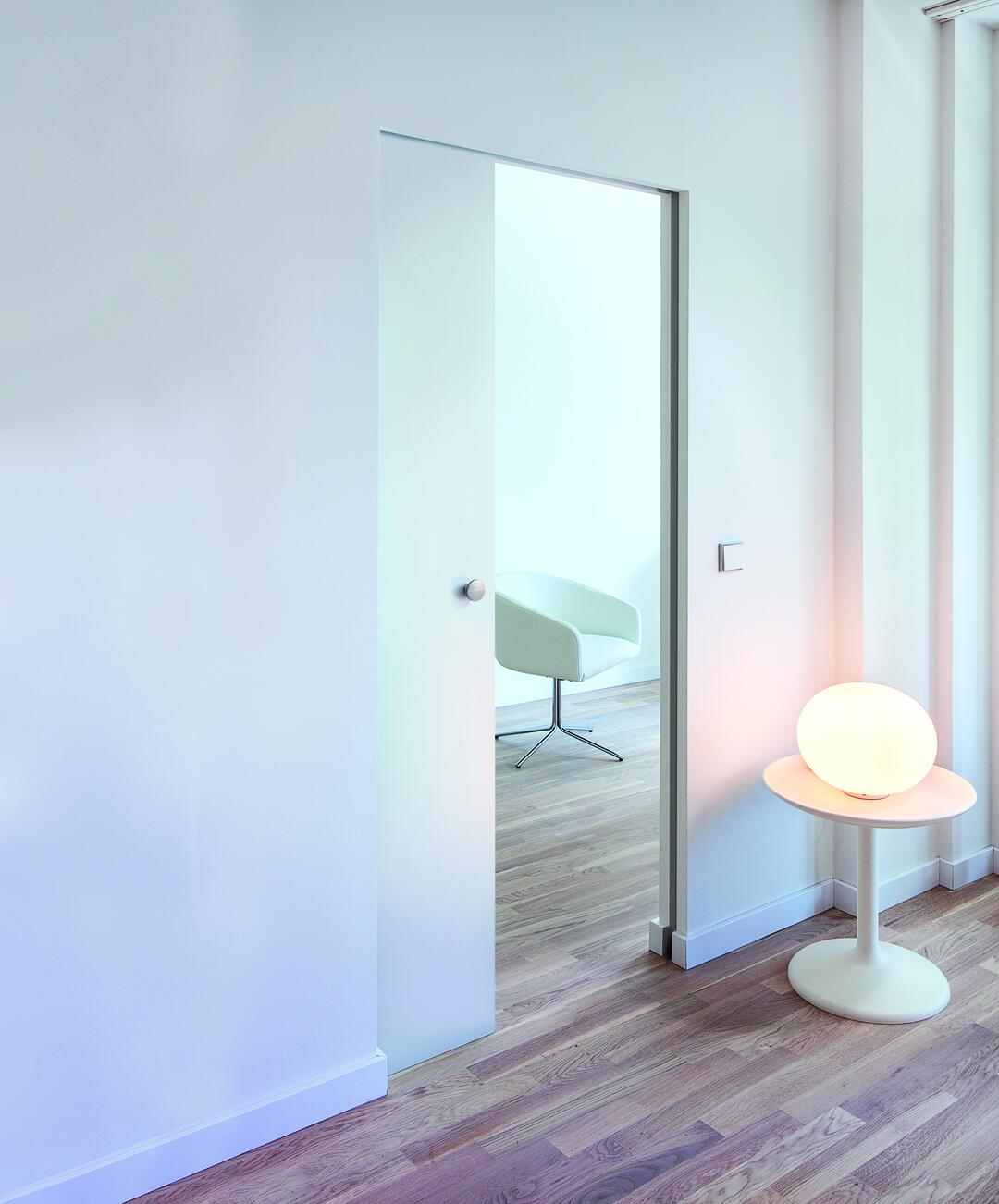 Blick auf eine weiße Wand mit einer weißen Knauf-Schiebetür, dahinter ein heller Raum, neben der Tür ein Beistelltisch mit einer leuchtenden Lampe.
