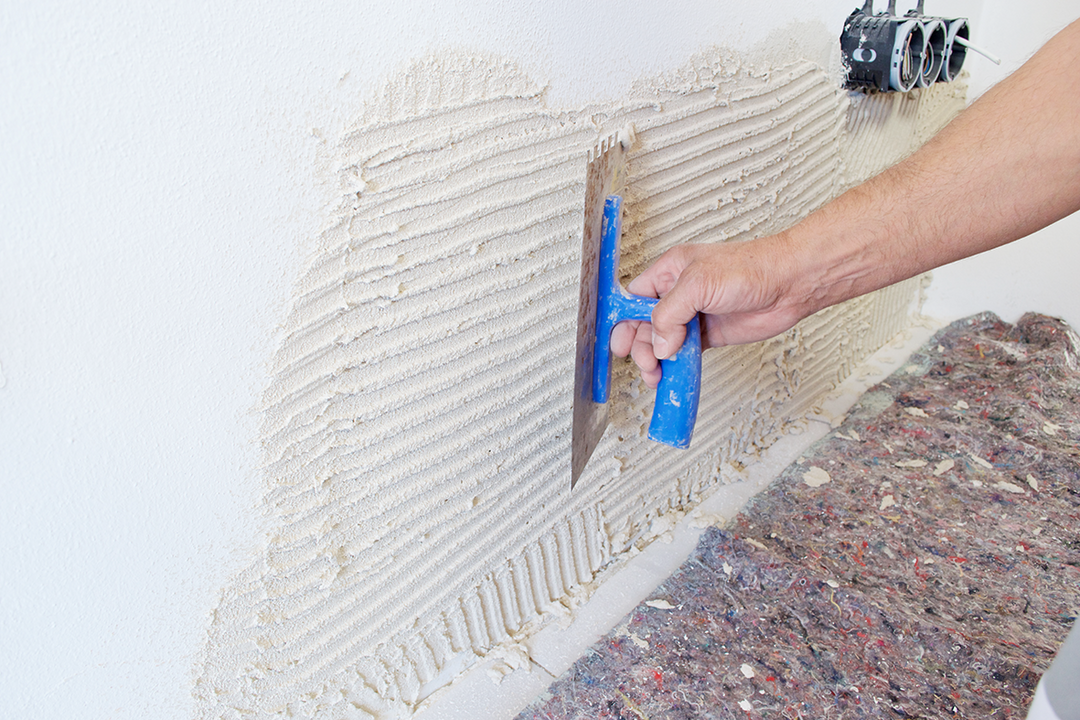 Eine Hand verteilt mit einem blauen Gerät eine Haftmasse auf einer weißen Wand. Der Boden ist mit Malervlies geschützt.