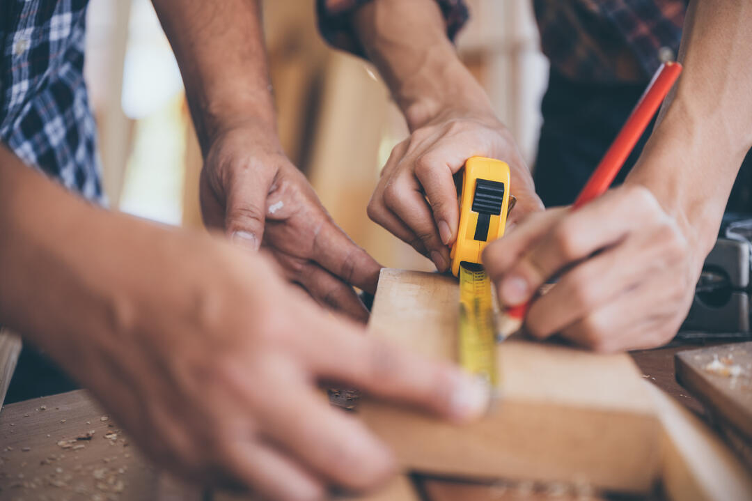 Zwei Paar Hände arbeiten an einem Stück Holz. Eine Person hält Lineal und Bleistift in der Hand.