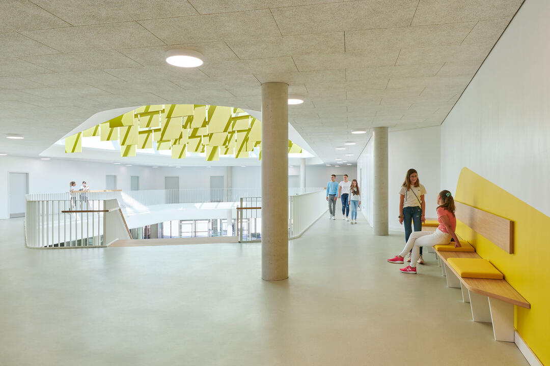 Ein heller, einladender Flur in einer Schule, mit einer texturierten Decke, gelben Sitzbänken und Säulen, die die Decke halten.