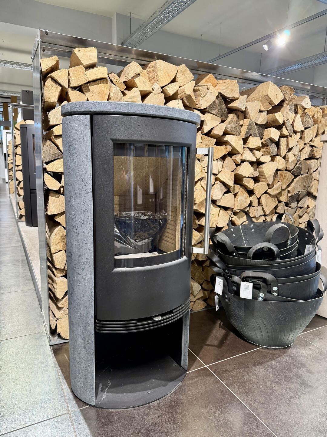 Ein moderner schwarzer Kaminofen in einem Geschäft vor einem großen Holzstapel. Der Ofen wirkt bullig und robust. Er ist schwarz, hat ein Holzfach und eine große Glasscheibe.