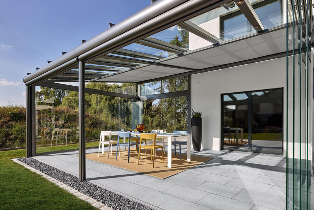 Ambientebild von einer Terrasse aus LuxoGran-Terrassenplatten in der Farbvariante Platingrau vor einem modernen weißen Gebäude unter blauem Himmel.