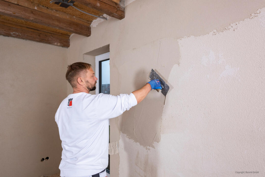Ein junger Handwerker in einem weißen Pullover mit dem baumit-Logo arbeitet in einem Innenraum. Er bringt die Produktmasse auf die Wand auf.