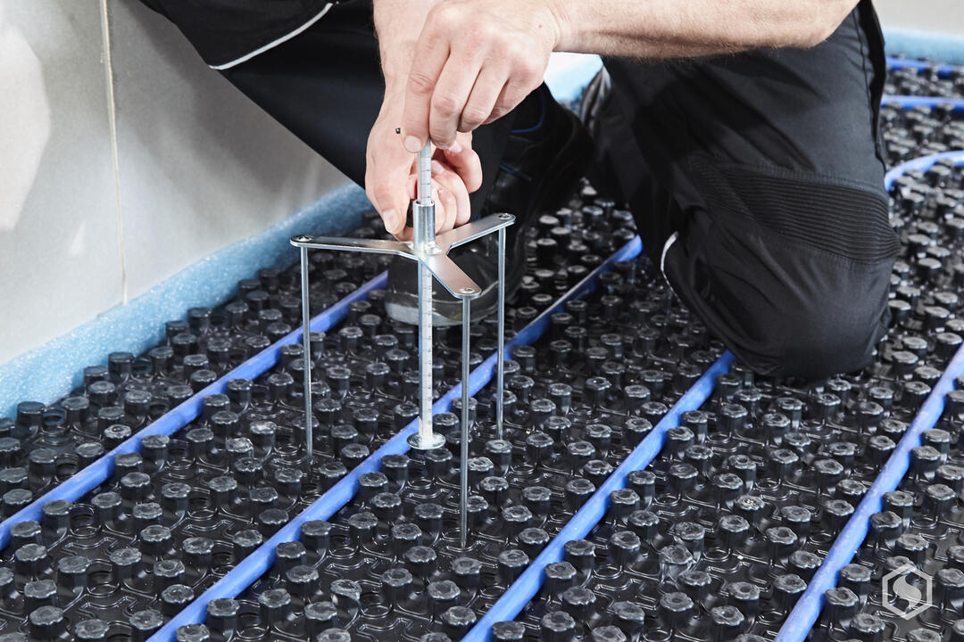 Ein Handwerker, von dem man nur den hockenden Unterkörper sieht, bestimmt die Höhe der Spachtelmasse mit einer Estrichspindel.