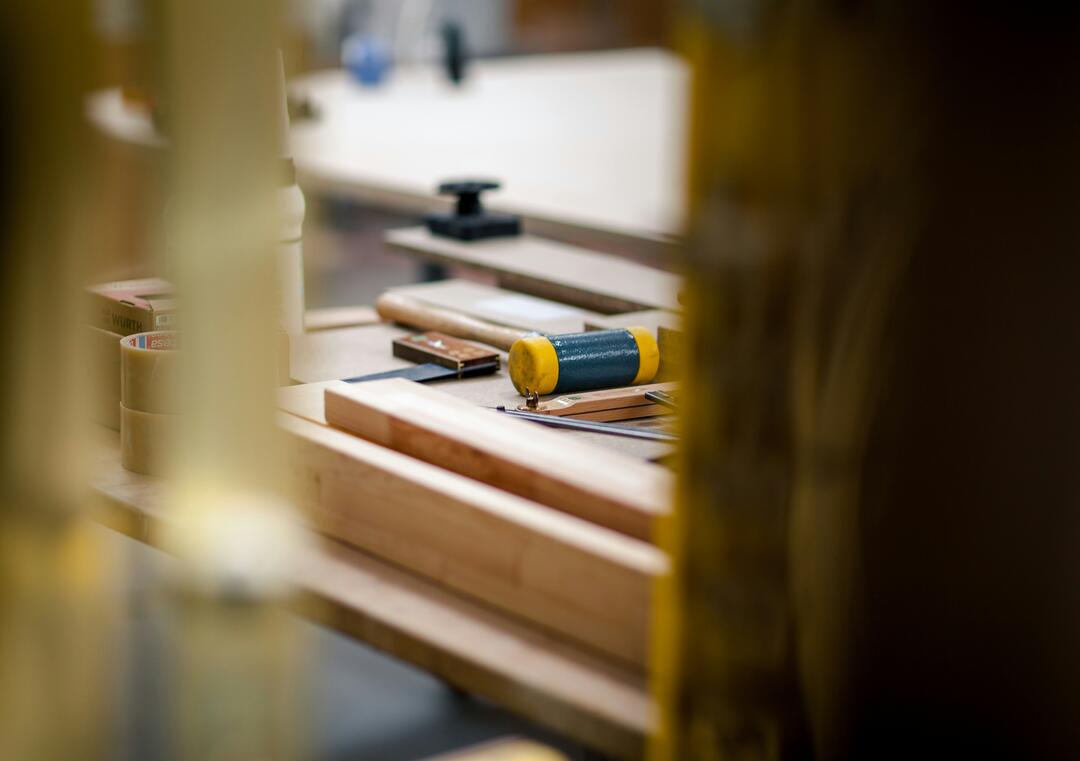 Werkzeuge auf einer hölzernen Werkbank, fotografiert durch die Streben eines Holzregals. Die Werkzeuge haben JC-Farben, also Gelb und Blau.