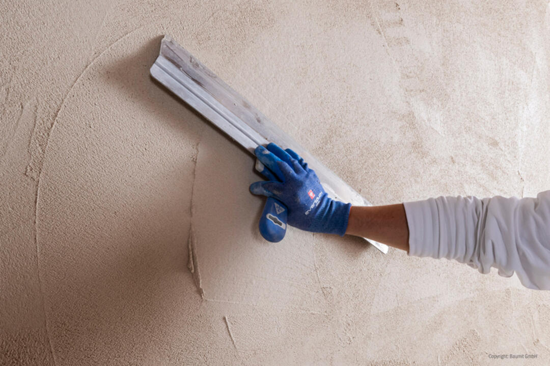 Eine Hand mit einem blauen Handschuh bringt mit einem Spachtel die Produktmasse auf eine Wand auf und verteilt sie gleichmäßig.