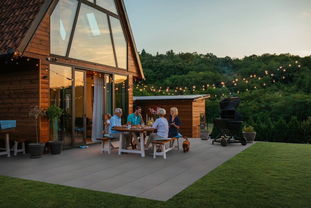 Ambientebild von einer Terrasse aus LuxoGran-Terrassenplatten in der Farbvariante Dunkelgrau vor einem Blockhaus mit großen Glasfenstern und viel Grün im Hintergrund. Es herrscht weiche Abendstimmung. Personen sitzen auf der Terrasse und essen etwas.