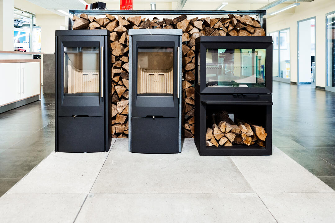 Drei verschiedene schwarze Kaminöfen vor einem Holzlager in einem modernen Ausstellungsraum.