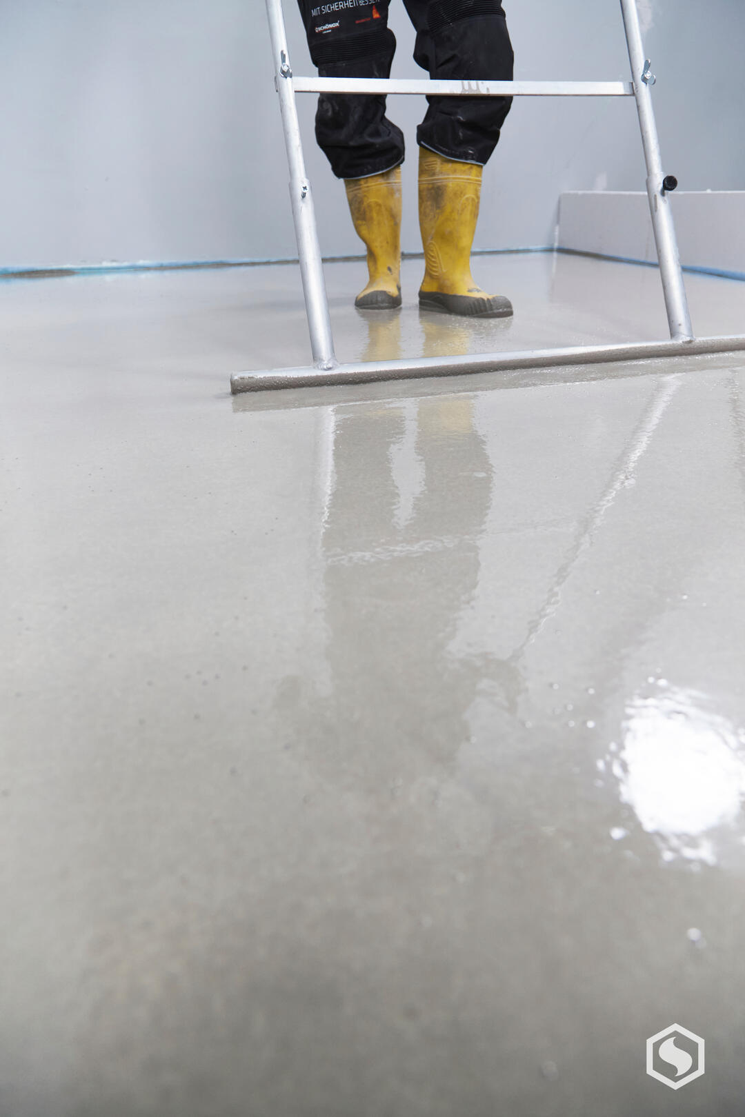 Ein Handwerker, von dem man nur die Füße in gelben Gummistiefeln sieht, bearbeitet eine Bodenoberfläche mit einer Schwabbelstange.