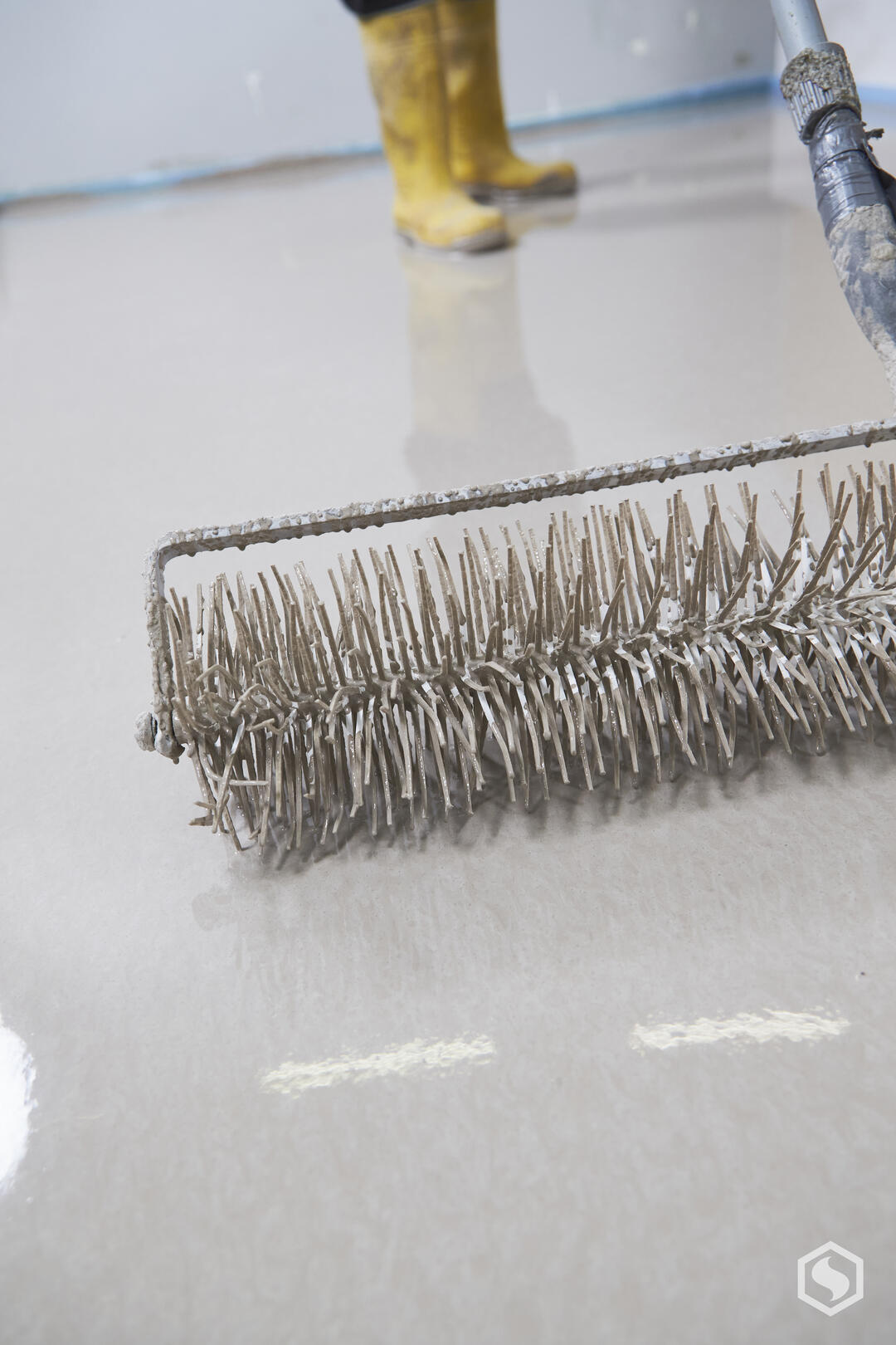 Eine Stachelwalze, mit der ein Bodenbelag entlüftet wird, in der Detailaufnahme. Im Hintergrund der gelbe Gummistiefel eines Handwerkers.