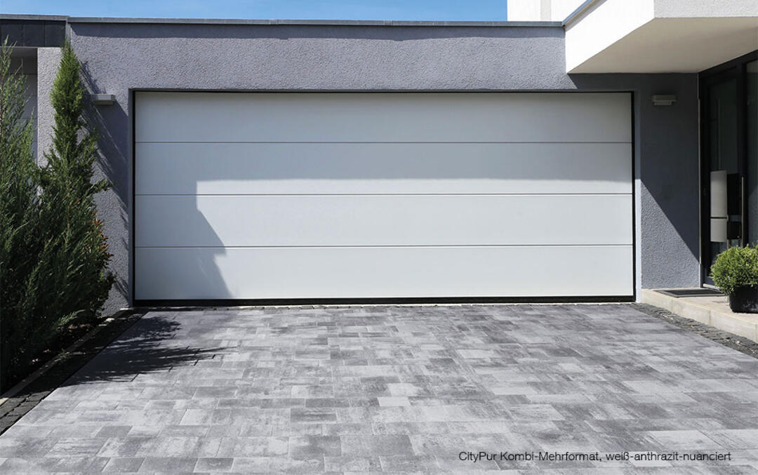 Eine Garage mit breitem, weißem Tor, die an ein Gebäude anschließt. Dahinter blauer Himmel, davor eine gepflasterte Auffahrt, deren vielfältiges Verlegebild einen interessanten Kontrast zum modernen Look der Garage bildet. Grünpflanzen flankieren die Auffahrt.