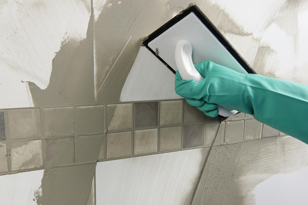 Eine Hand in einem grünen Putzhandschuh bewegt einen Spachtel über eine mit Fugenmörtel verschmierte Wand und verteilt so den Mörtel in alle Ritzen.