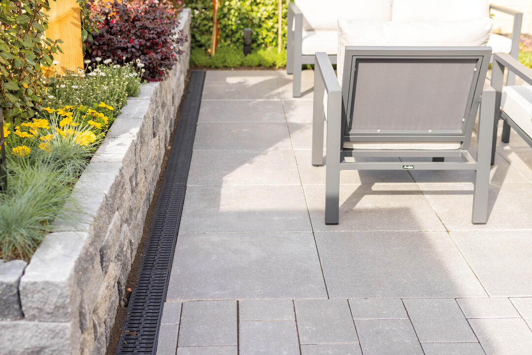 Eine mit Steinzeug geflieste Terrasse, auf der eine Sitzgruppe steht. Eine Steinmauer grenzt an, hinter der sich Pflanzen anschließen. Zwischen der Mauer und der Terrasse schmiegt sich ein längliches, schwarzes Regenwasserableitungselement von ACO in den Boden.