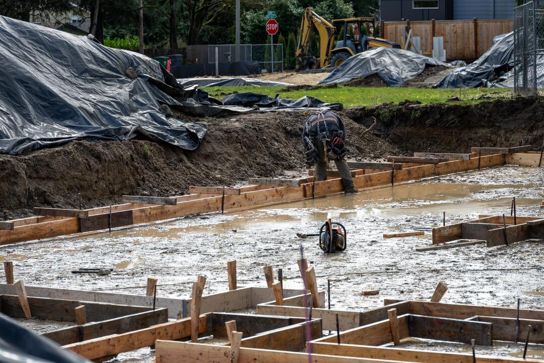 Eine Baustelle mit offenliegendem Fundament eines Hauses bei Regen, der sandige Boden ist matschig und ein Arbeiter bückt sich im Hintergrund nach etwas.