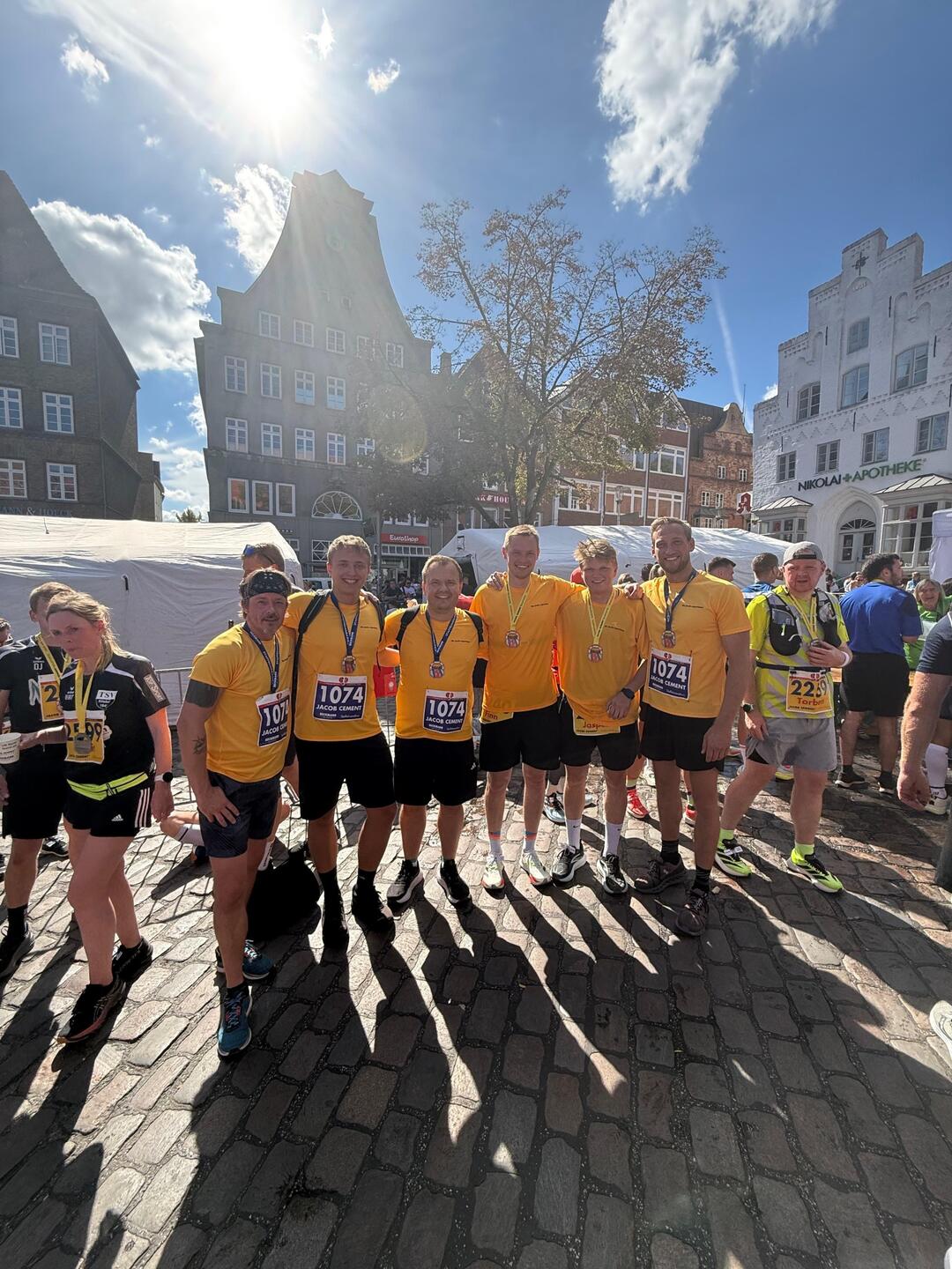 Die JACOB CEMENT-Mannschaft posiert in gelben Unternehmenstrikots für ein Gruppenfoto auf dem Flensburger Südermarkt. Die Sonne scheint und die Menschen auf dem Bild lächeln.
