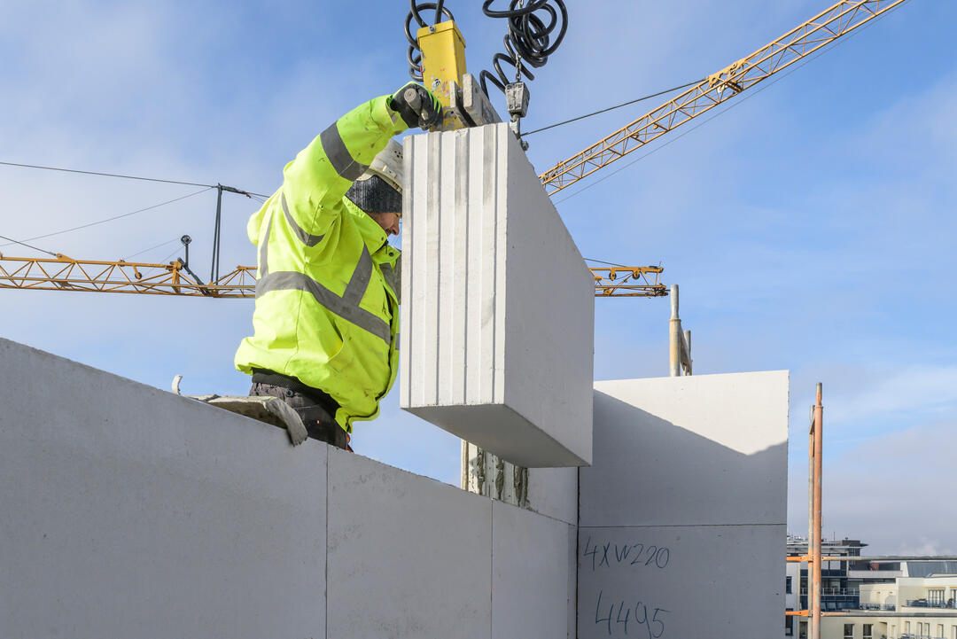 Ambientebild von der Baustelle; ein Bauarbeiter in Schutzjacke zieht ein Silka Protect XL-Basic-Element an einem Kran zu sich herunter und auf eine in Konstruktion befindliche Mauer. Dahinter blauer Himmel und ein großer gelber Kran.