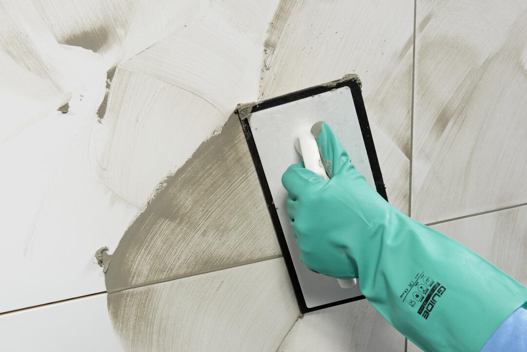 Eine Hand in einem grünen Putzhandschuh bewegt einen Spachtel über eine mit Fugenmörtel verschmierte Wand und verteilt so den Mörtel in alle Ritzen.
