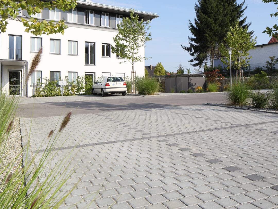 Aufgeweitete Fugen wie auf diesem gepflasterten Platz eignen sich sehr gut für versickerungsfähige Flächen. Im Hintergrund sind ein weißes Haus, Büsche und Bäume zu sehen.