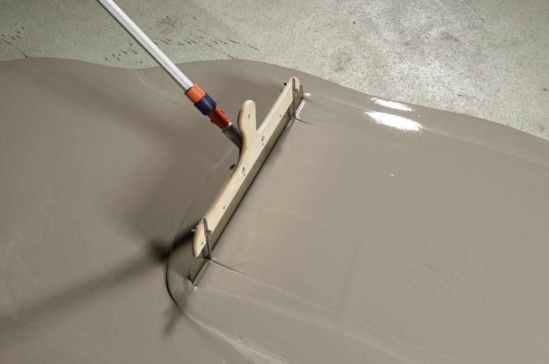ARDEX K 15 N-Spachtelmasse wird auf dem Boden verteilt. Sie hat einen satt-warmen, hellgrauen Farbton und glänzt im Licht.