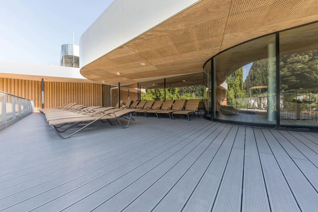 Ein großzügiger Außenbereich einer Wellnesseinrichtung mit Terrassendielen in Holzoptik am Boden und vielen Liegestühlen. Das Gebäude dahinter ist rund mit einer großen Glasfront und einem überhängenden flachen Dach.