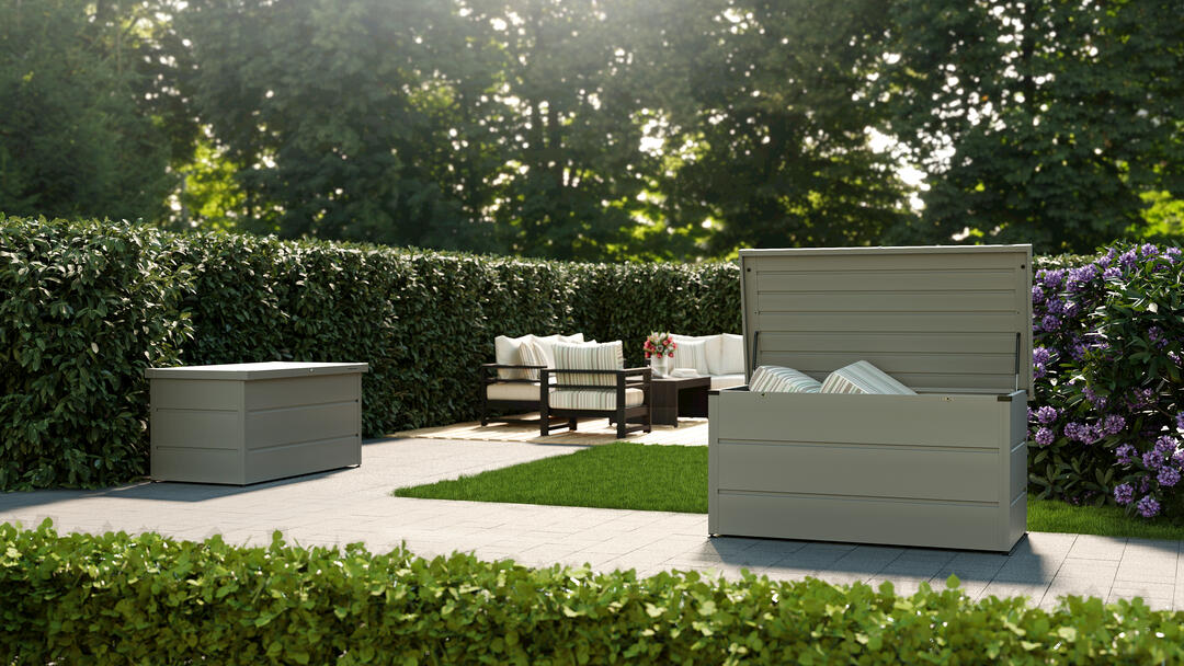 Eine Terrasse, umgeben von Bäumen und grünen Hecken, im Vordergrund eine aufgeklappte Gartenbox von Hörmann, hell und geräumig.