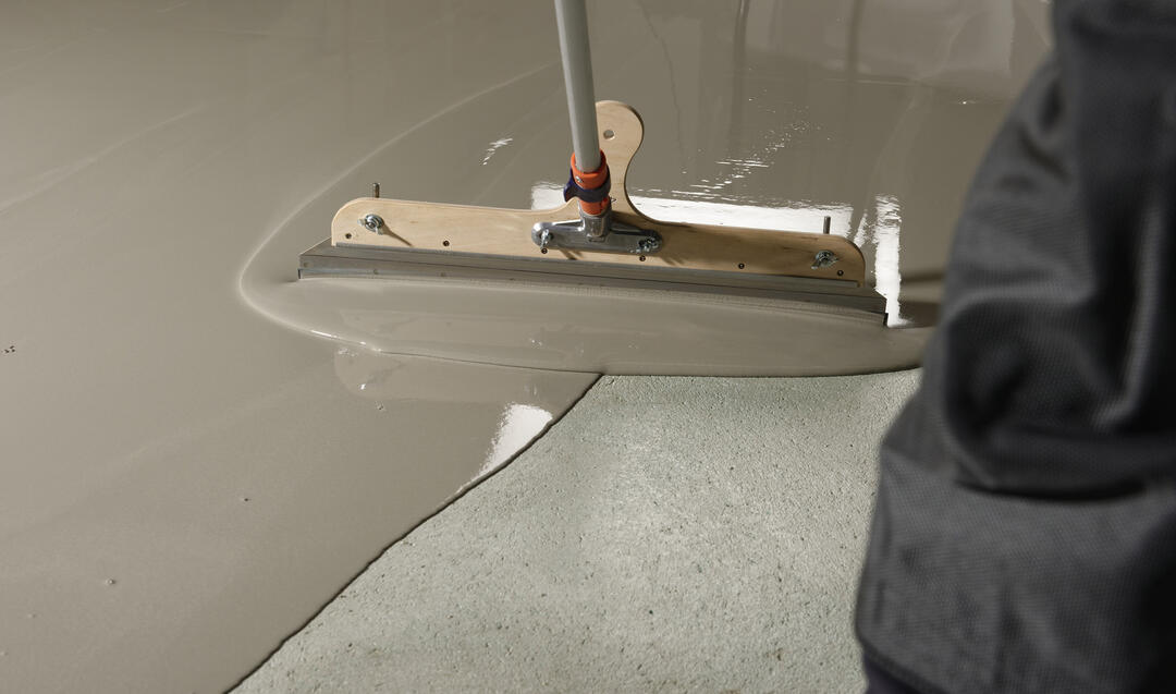 ARDEX K 15 N-Spachtelmasse wird auf dem Boden verteilt. Sie hat einen satt-warmen, hellgrauen Farbton und glänzt im Licht.