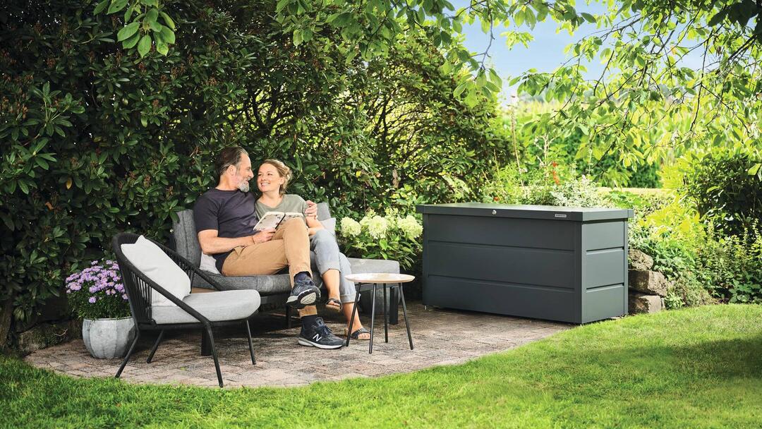 Ein Paar sitzt in einem grün blühenden Garten auf einer Sitzgelegenheit und lächelt sich an. Daneben steht eine Gerätebox von Hörmann.