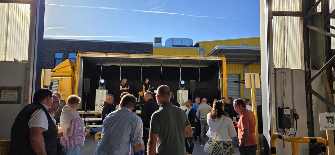 Bei einer ROCK-AM-REGAL-Veranstaltung von JACOB CEMENT stehen Menschen vor einer Bühne unter freiem Himmel. Das Bild ist aus einer Lagerhalle heraus fotografiert.