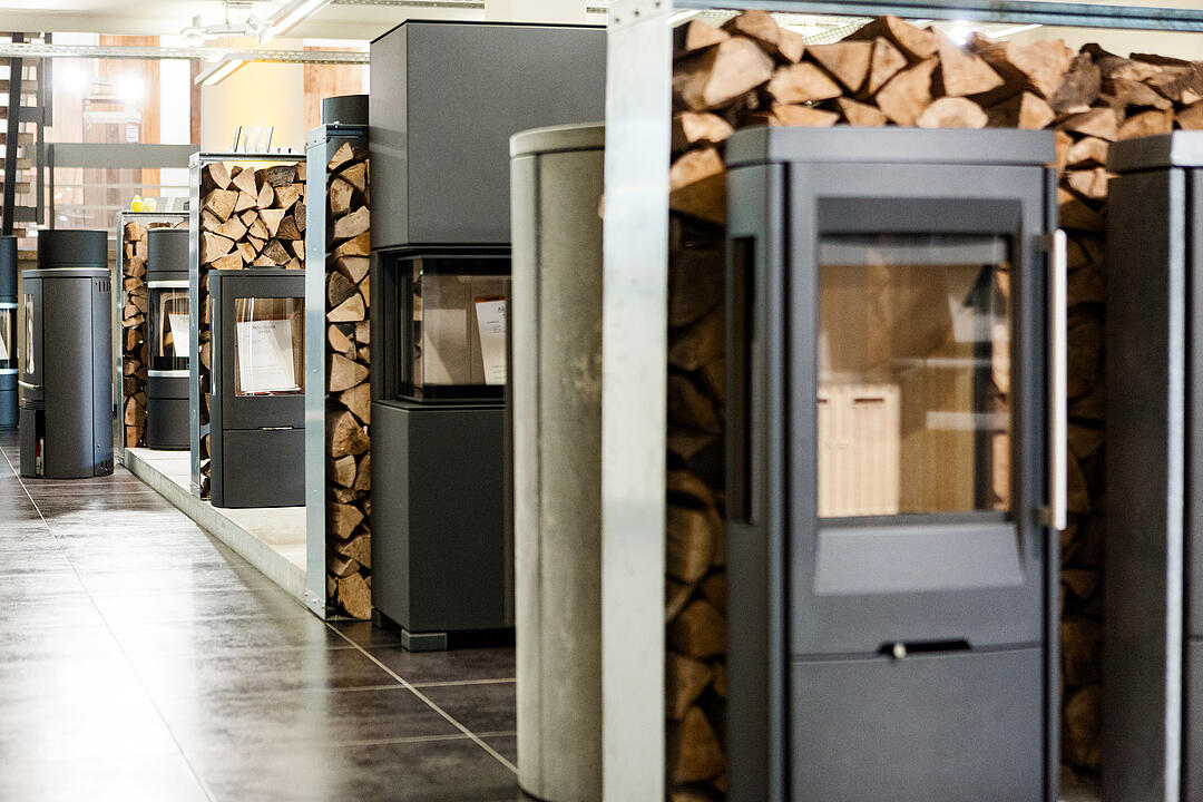 Fünf Kamine hintereinander aufgereiht, jeweils mit gestapelten Holzscheiten in der Ausstellung von JACOB CEMENT BAUSTOFFE